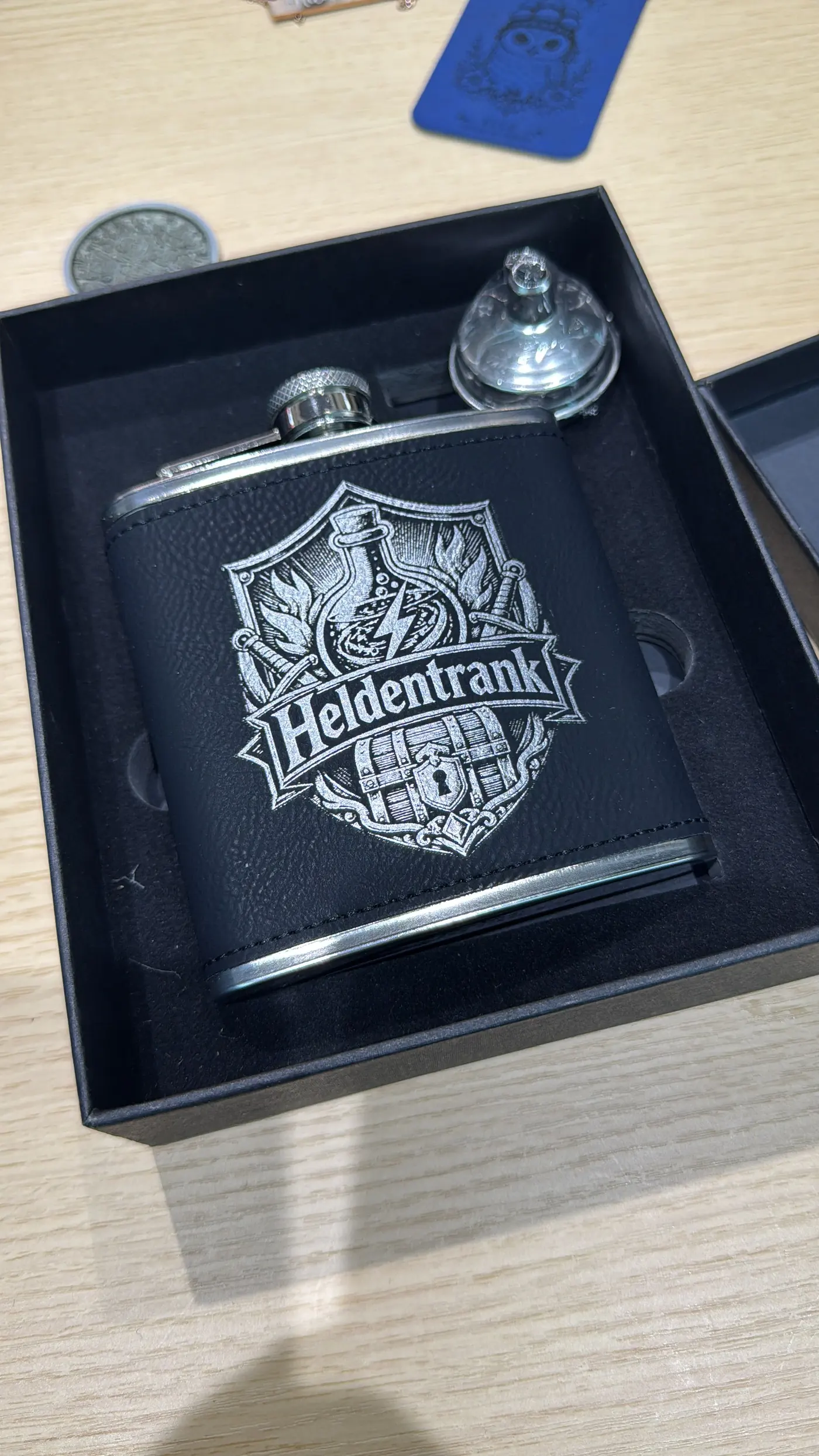 Ein schwarzer Flachmann mit Kunstlederbezug liegt in seiner offenen Geschenkschachtel, graviert mit einem Wappen und dem Schriftzug „Heldentrank“; daneben liegt ein kleiner Trichter.