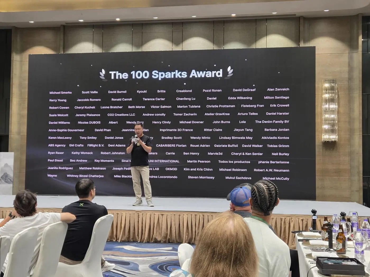 Ein Redner mit Mikrofon auf einer Bühne in einem Festsaal, hinter ihm eine große Leinwand mit der Überschrift „The 100 Sparks Award“ und darunter rund hundert Namen in Spalten, unter ihnen „Key Moments“; im Vordergrund Gäste an gedeckten Tischen, von hinten zu sehen.