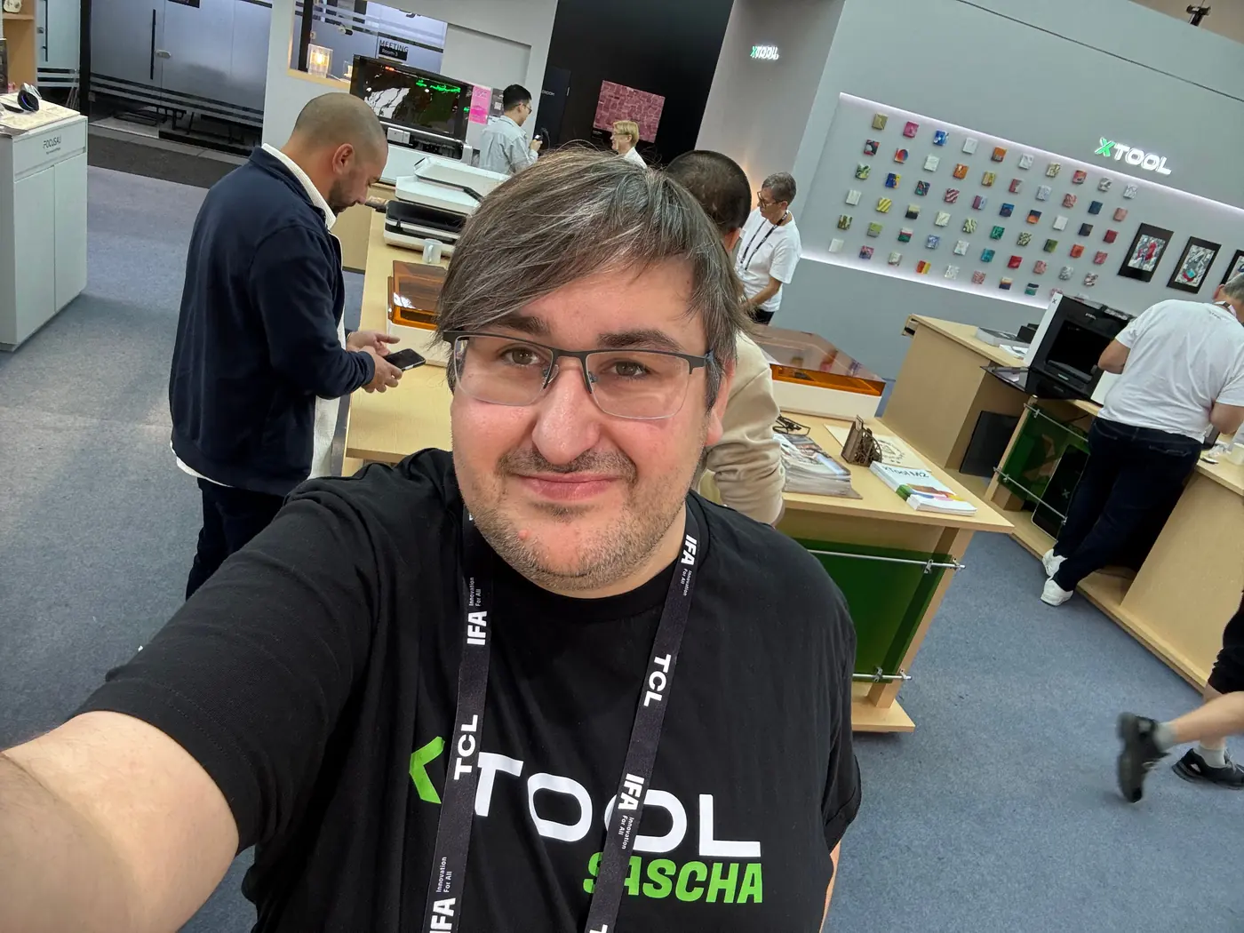 Sascha Pfeuffer im schwarzen xTool T-Shirt mit IFA-Lanyard am Messestand von xTool auf der IFA 2026 in Berlin, im Hintergrund eine Wand mit bunten Materialproben und Arbeitsplätze mit Geräten.