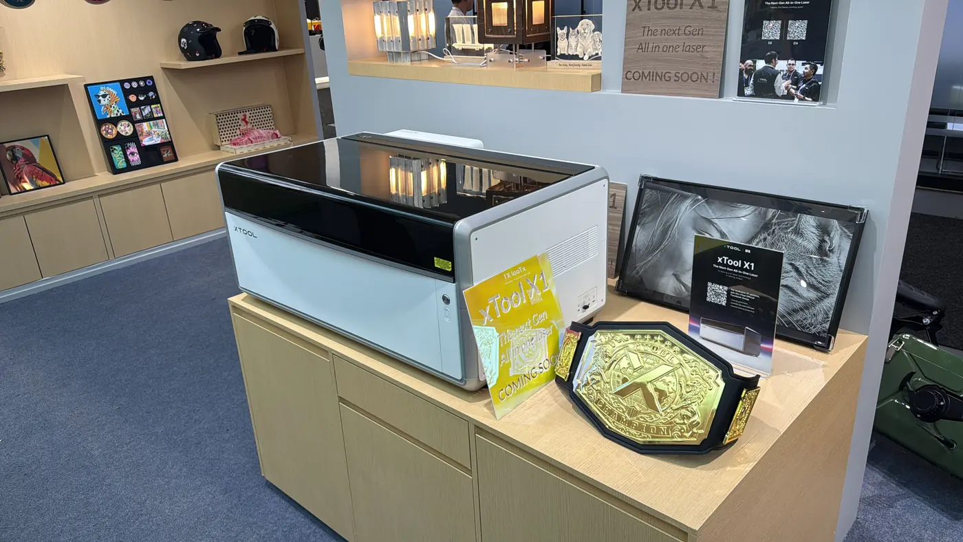 Ein weiß-schwarzer xTool X1 steht auf einem hölzernen Podest am Messestand, daneben ein gelbes Acrylschild „xTool X1“, eine gravierte schwarze Platte und ein goldener Champions-Gürtel; an der Wand dahinter ein Plakat mit der Zeile „The next Gen All in one laser – Coming soon“.