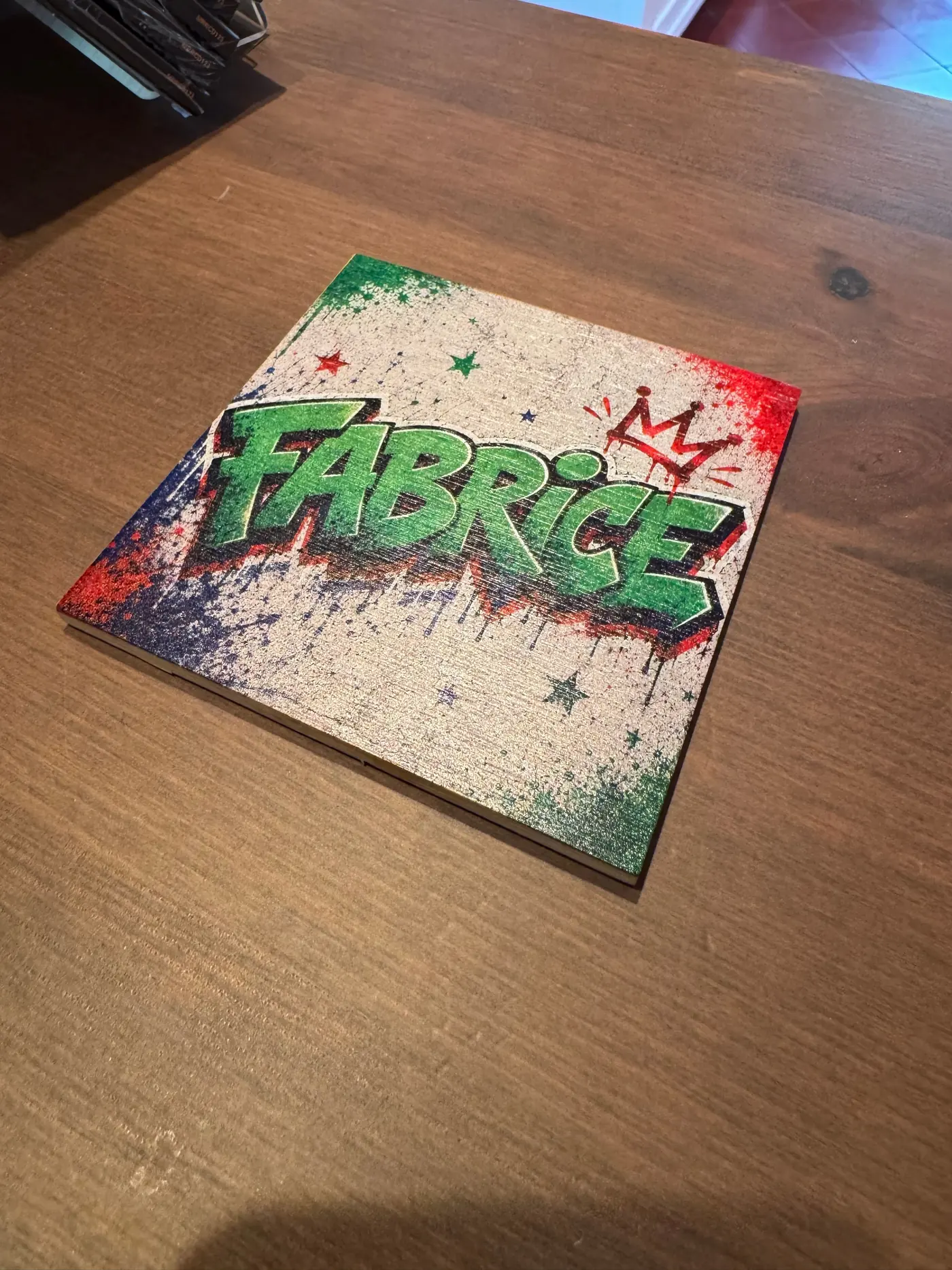 A square bamboo coaster on a dark wooden table, printed with the graffiti lettering “Fabrice” in green with a red crown on a speckled, colourful ground.