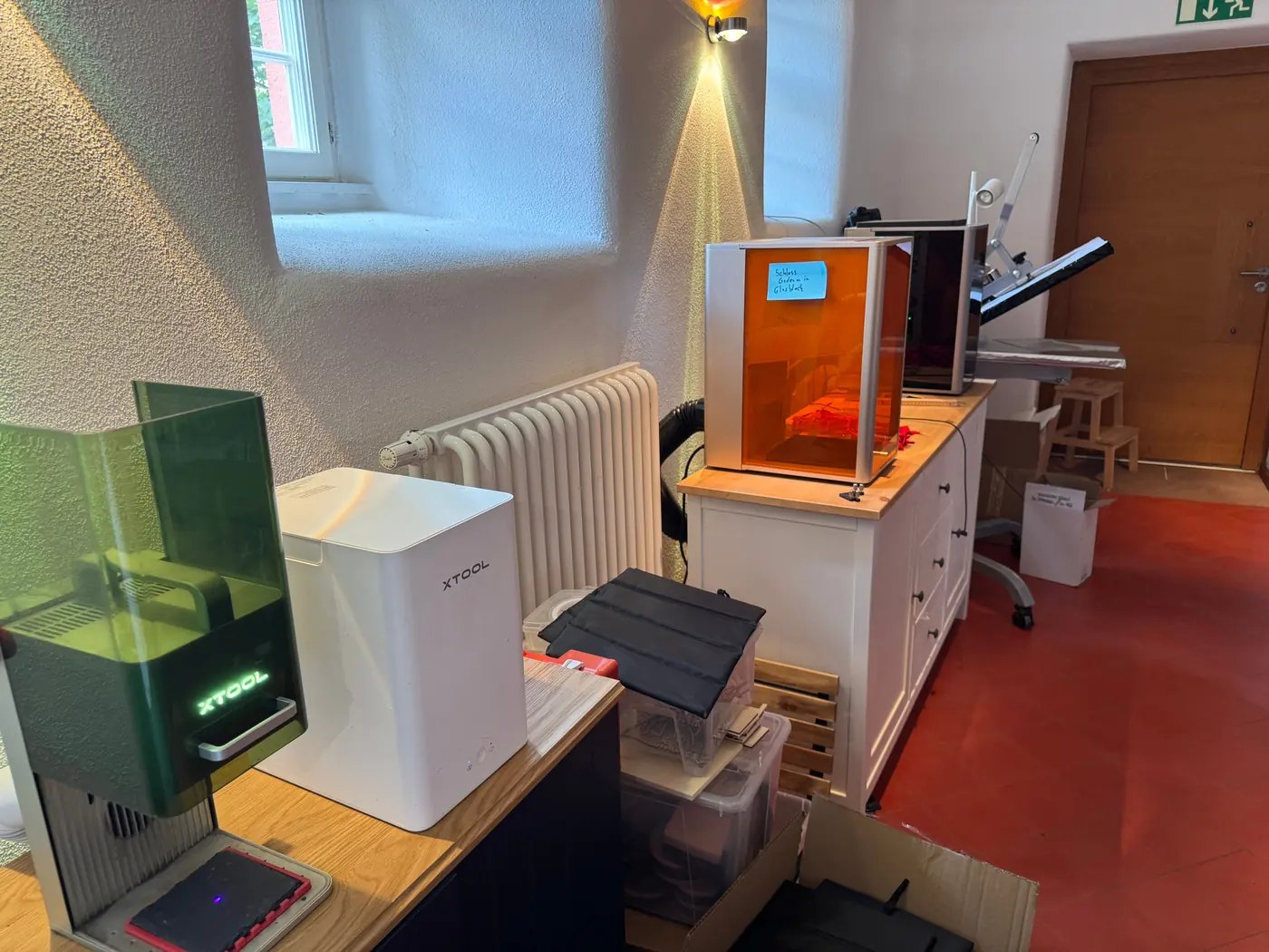 Blick in den Demo-Raum in Gedern: links auf einem Sideboard der xTool F1 mit weißem Rauchfilter, im Hintergrund der xTool F2 Ultra UV mit orangefarbener Scheibe, daneben eine Transferpresse.