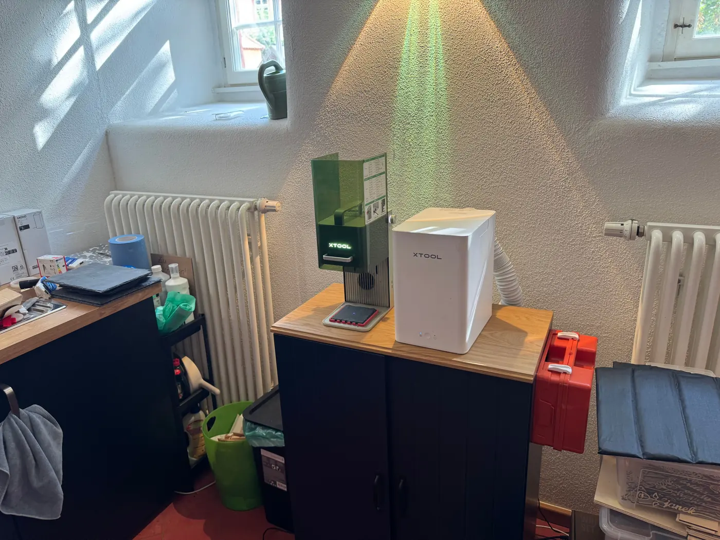 The Gedern work spot from further back: the xTool F1 and the fume filter on a dark sideboard between two radiators, a workbench with material on the left.
