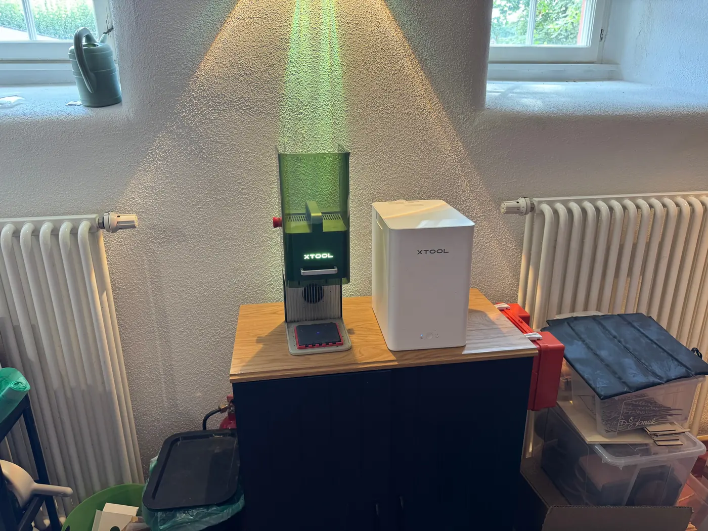 Der xTool F1 mit grüner Schutzhaube steht neben einem weißen Rauchfilter auf einer hellen Holzarbeitsplatte, links und rechts je ein Heizkörper.