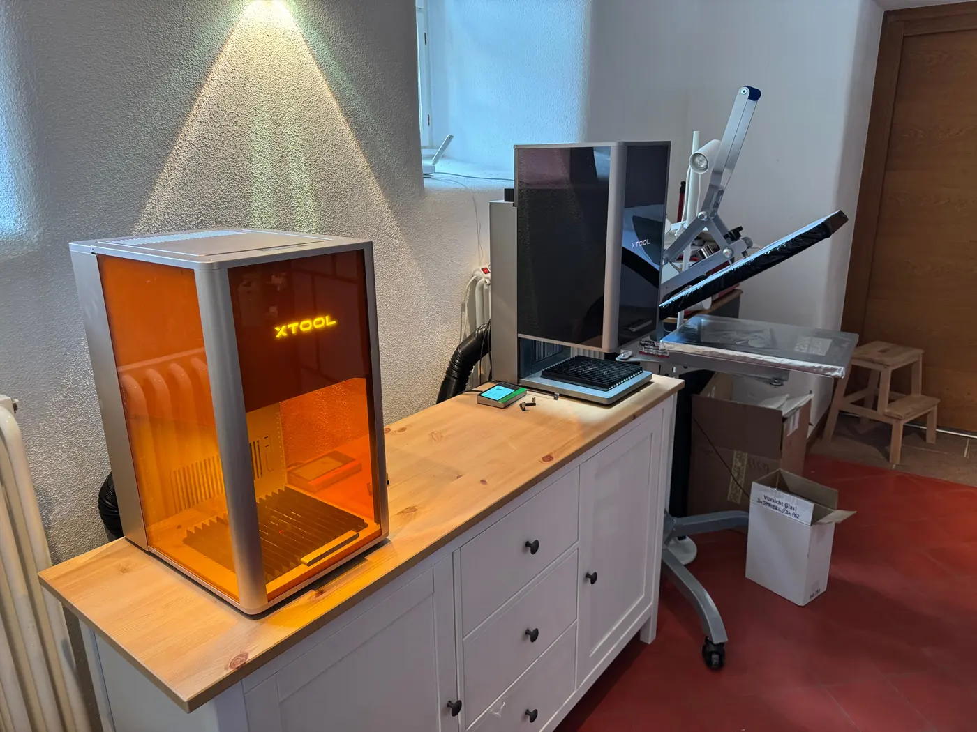 Der xTool F2 Ultra UV mit orangefarbener Schutzscheibe steht vorn auf einem Sideboard, dahinter der dunkle xTool F2 Ultra, rechts eine Transferpresse.