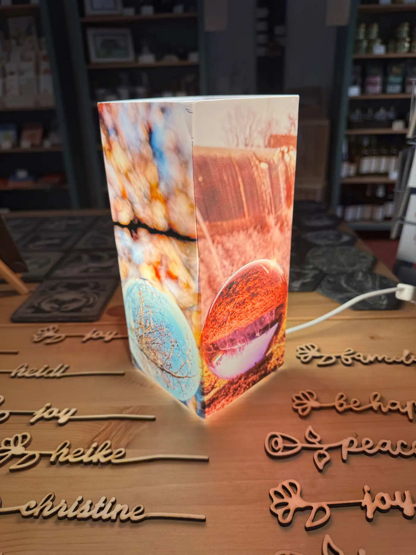 A lit Gröno table lamp, printed all round with photographic motifs, standing on a counter among laser-cut wooden first names.