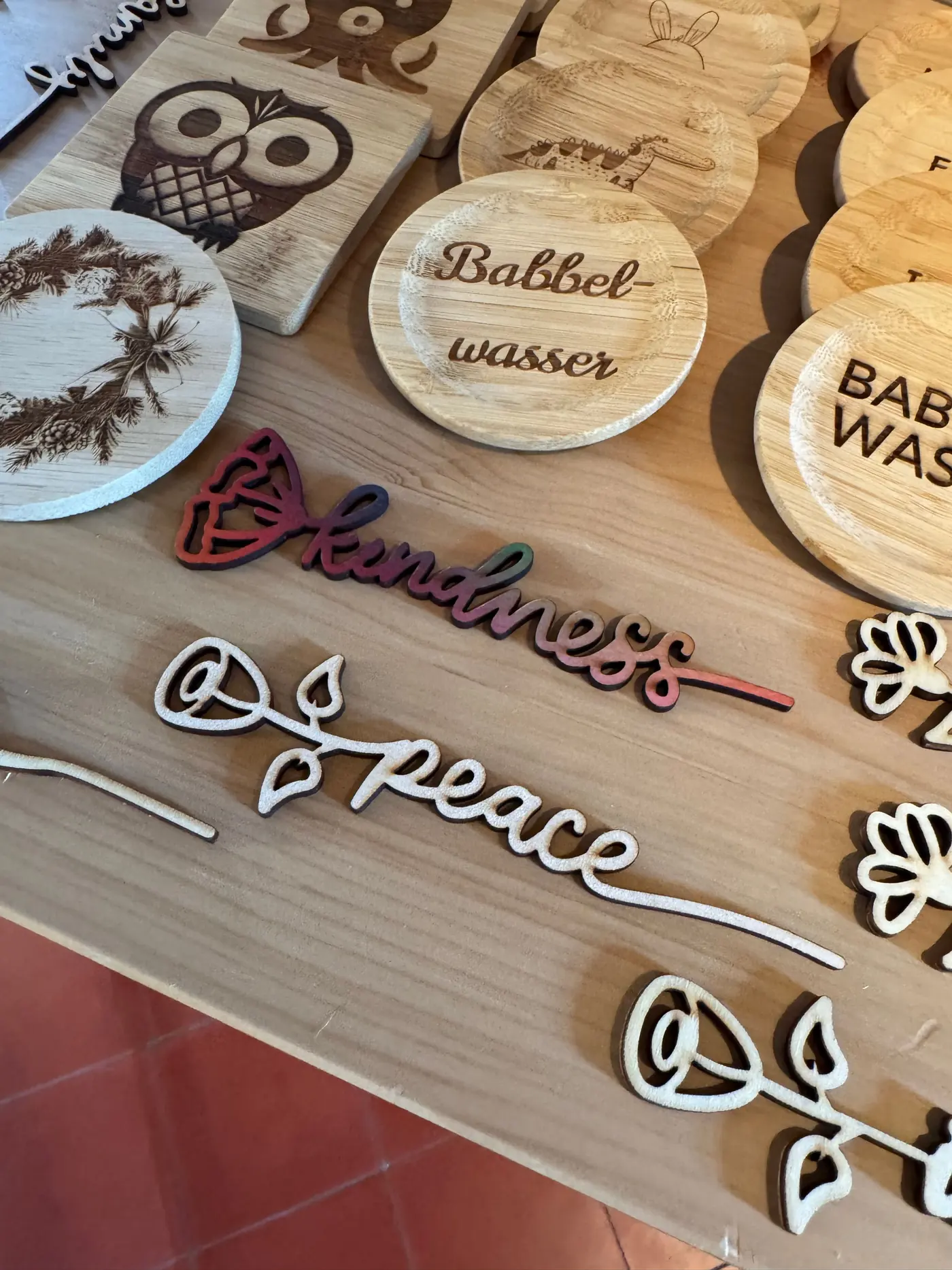 Laser-cut plywood lettering on a pale wooden table, one piece at the front with a blossom and a colour gradient, unprinted pieces and engraved bamboo coasters beside it.