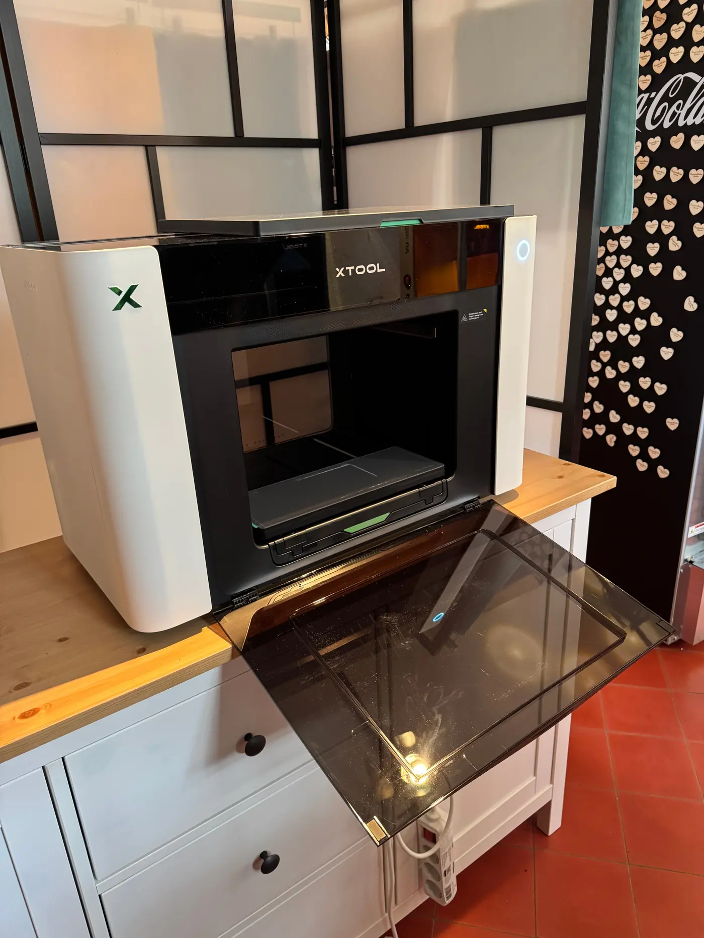 Der xTool O1 Omni Printer steht im Ladenraum auf einer Kommode, die Glasklappe ist heruntergeklappt, dahinter eine Glastrennwand.