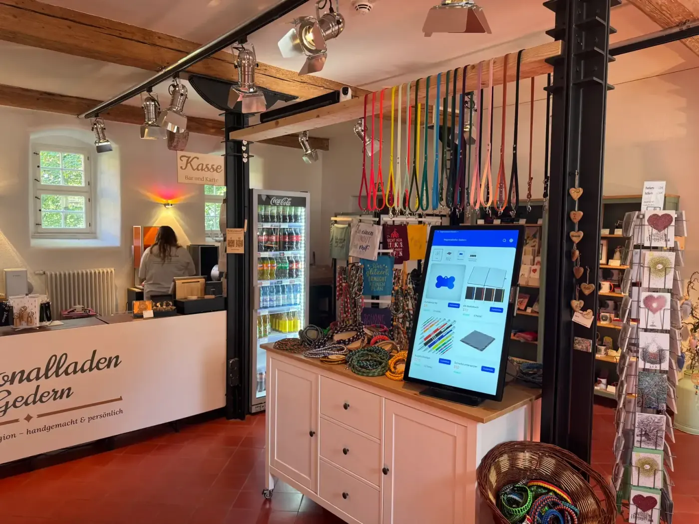 Der xTool Self-Service Kiosk steht hochkant auf einem Sideboard im Regionalladen Gedern, auf dem Bildschirm der Katalog mit Hundemarke, Notizbuch, Kugelschreiber und Schieferuntersetzer. Dahinter die Kasse, darüber hängen Schlüsselbänder.